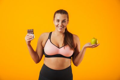 Imagen de feliz mujer con sobrepeso en chándal sosteniendo barra de chocolate y manzana en ambas manos, aislado sobre fondo amarillo